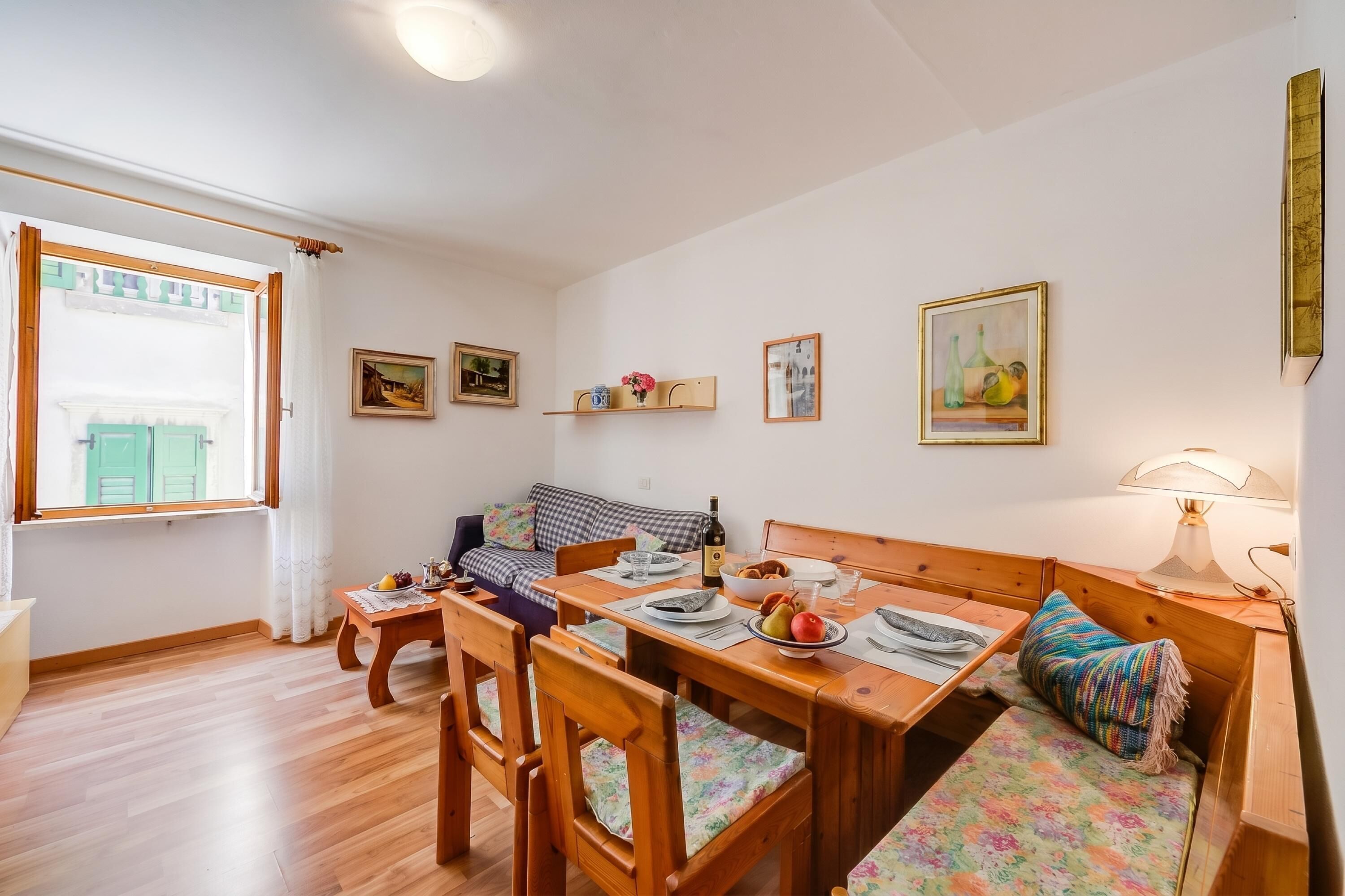 A cozy room with a wooden dining table, chairs, a bench, and a sofa. There are framed pictures on the walls, a window with curtains, and a lamp on the bench.