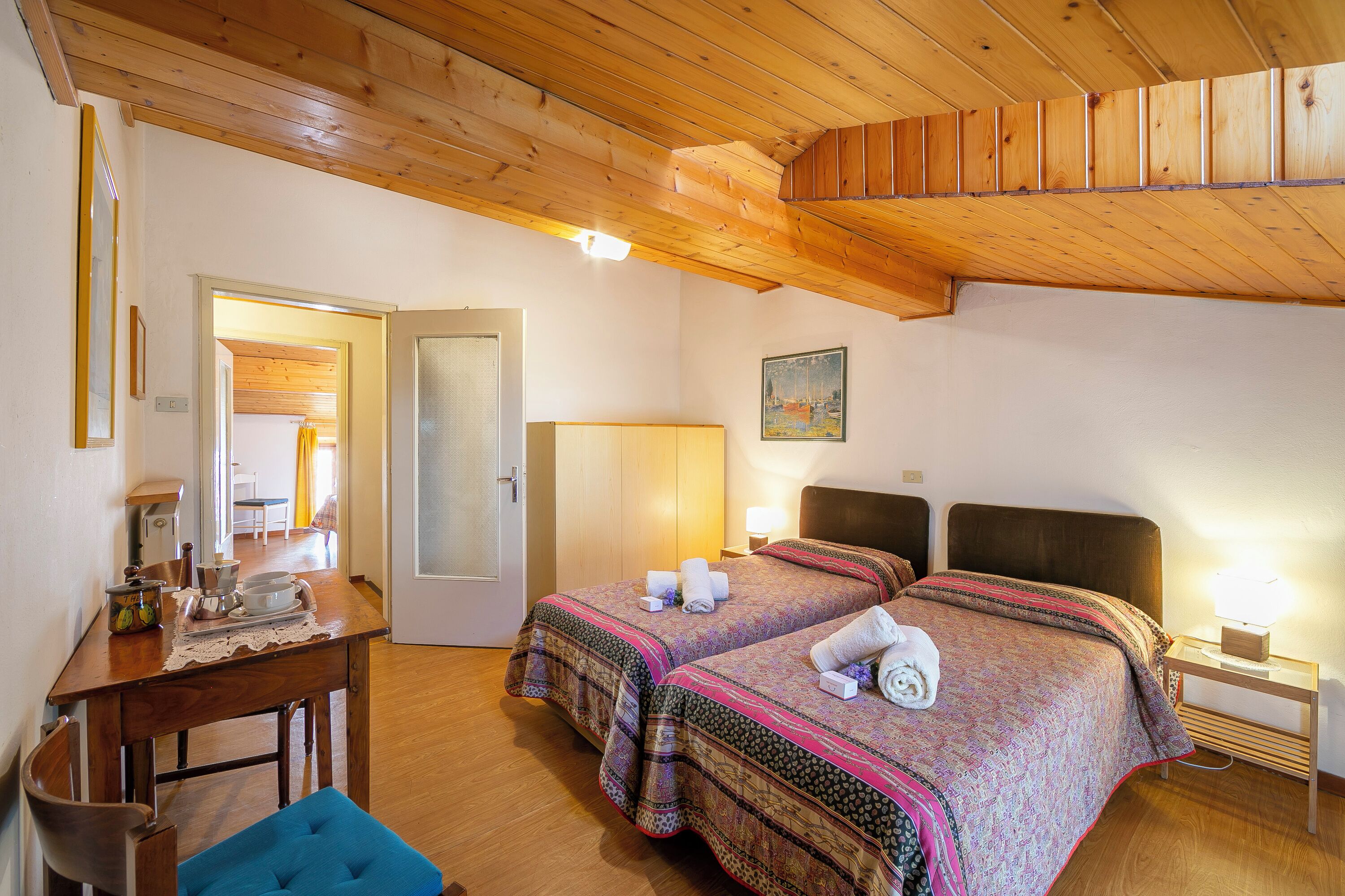 A room with two beds, a wooden ceiling, a desk with a chair, and a small table with a lamp.