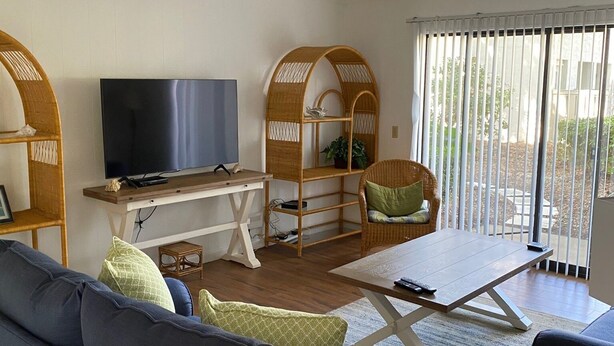 A living room with a sofa, a TV, a coffee table, and a wicker chair. - Surf Court Unit #1-Remodeled villa across the street from the ocean (Hilton Head Island)