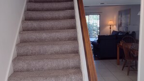 A carpeted staircase with a wooden handrail leading to a landing. The landing area has a tiled floor and a dark-colored sofa. - Surf Court Unit #1-Remodeled villa across the street from the ocean (Hilton Head Island)