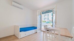 Una habitación con una cama blanca, una mesa de madera y una silla. Se ve una ventana con vistas a edificios.