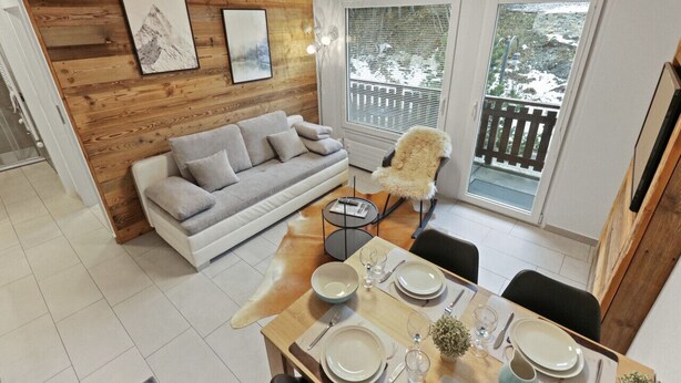 Una sala de estar moderna con una pared de madera, un sofá gris, una mesa de comedor preparada para cuatro personas y una vista de un paisaje nevado a través de amplios ventanales.