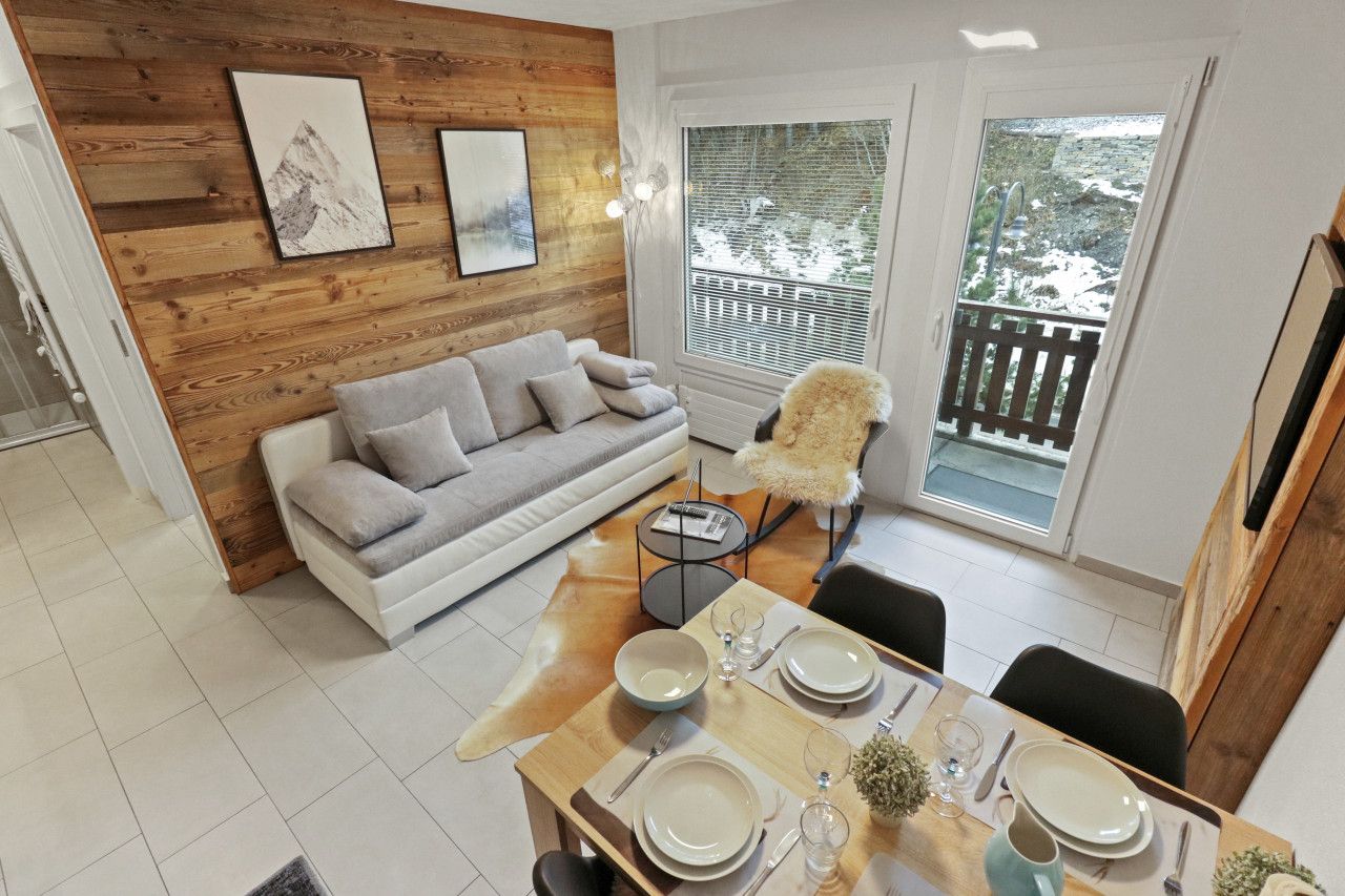Ein modernes Wohnzimmer mit einer Holzwand, einem grauen Sofa, einem Esstisch für vier Personen und Blick auf eine verschneite Landschaft durch große Fenster.