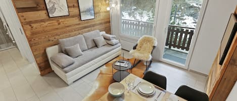 Een moderne woonkamer met een houten wand, een grijze bank, een eettafel gedekt voor vier personen en uitzicht op een besneeerd landschap door grote ramen.