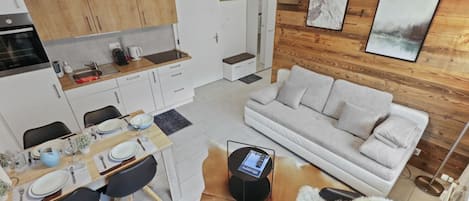 A modern kitchen with wooden cabinets, a dining area with a table set for four, and a living room with a sofa and a fluffy rug.