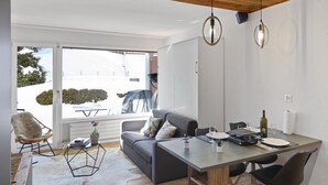 A modern living room with a grey sofa, a dining table with chairs, a wooden floor, and a large window with a view of a snowy landscape. - Studio rénové et élégant en plein centre (Crans-Montana)