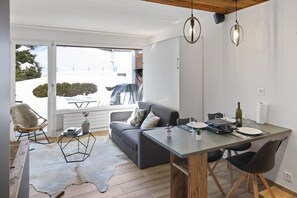 A modern living room with a grey sofa, a dining table with chairs, a wooden floor, and a large window with a view of a snowy landscape.