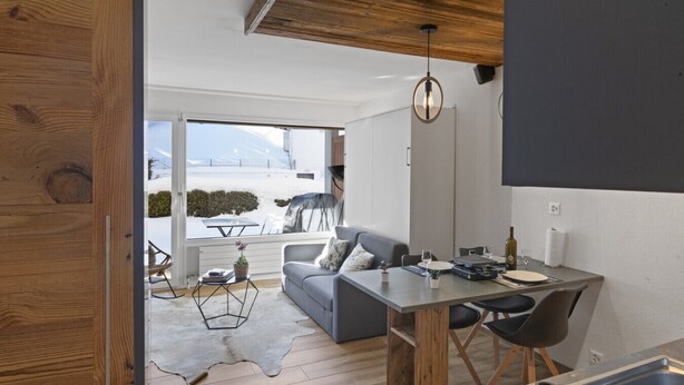 A modern kitchen with a dining area, a grey sofa, and a view of the outdoors. - Studio rénové et élégant en plein centre (Crans-Montana)