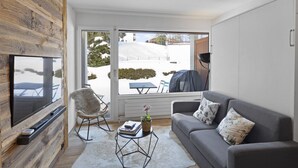 A modern living room with a TV, sofa, and chair, a large window with a view of a snowy landscape, and a wooden accent wall. - Studio rénové et élégant en plein centre (Crans-Montana)