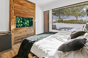 Een slaapkamer met een bed, een houten wandpaneel, een raam met uitzicht op een besneeerd landschap, en een klein tafeltje met stoelen erbuiten.