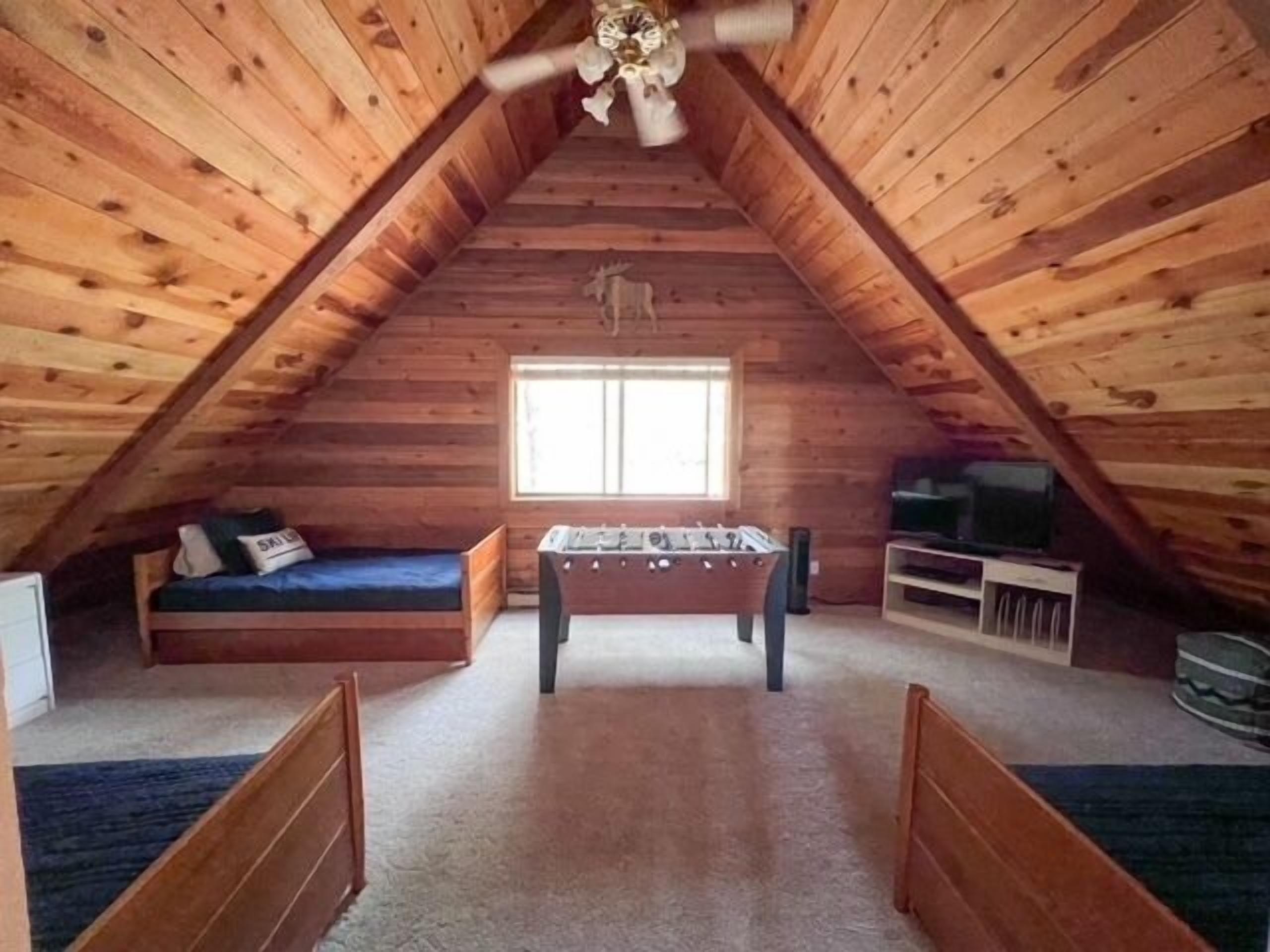A cozy attic room with wooden walls, a foosball table, a bed, and a TV.