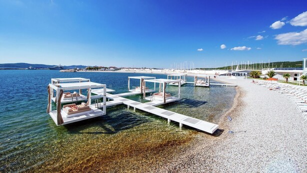 Une plage à l’eau cristalline, bordée de cabanas blanches et d’un port de plaisance où sont amarrés des bateaux.