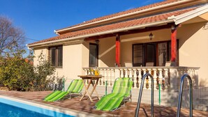 Un espace aménagé au bord de la piscine, avec deux chaises longues à rayures vertes, une petite table sur laquelle est posée une carafe, et un patio couvert doté d’une rambarde.