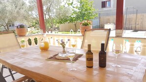 Une table en bois, avec deux bouteilles de vin et des verres, entourée de chaises, dans un cadre extérieur ensoleillé agrémenté de plantes en pot et offrant une vue sur un jardin.