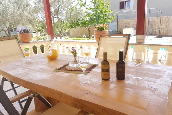 A wooden table with two wine bottles and glasses, surrounded by chairs, in a sunny outdoor setting with potted plants and a view of a backyard.