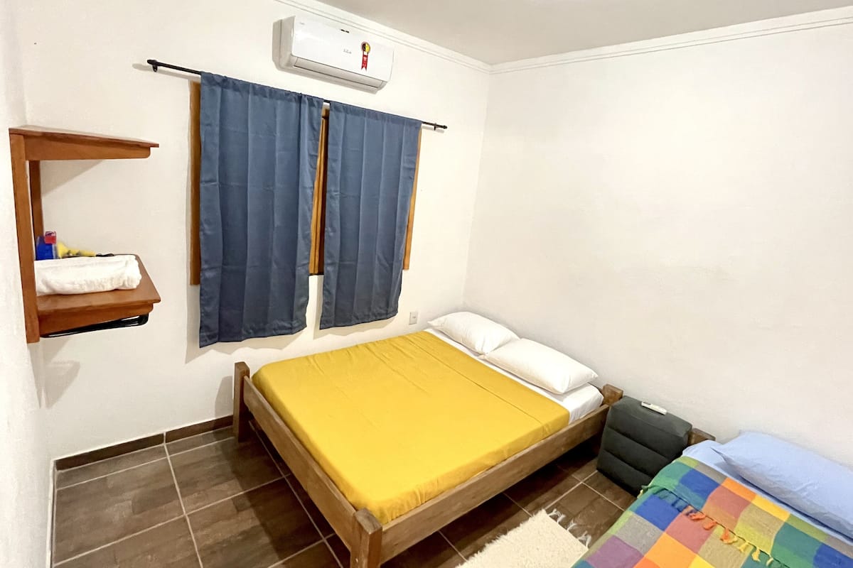 Quarto pequeno e limpo com cama, prateleira com produtos de higiene e janela com cortinas.