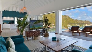 Amplia sala de estar con sofá, mesa de centro y televisor. Hay un balcón con sillas y vistas al paisaje.