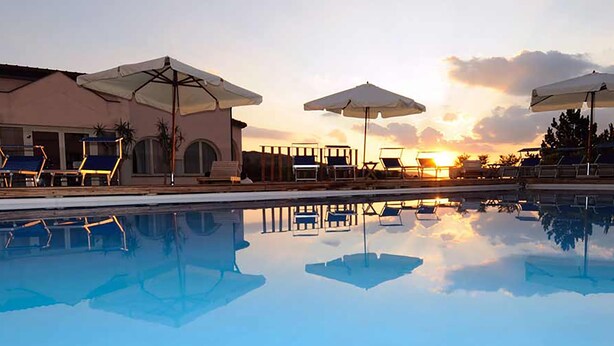 Un área de piscina con sillones reclinables y sombrillas, reflejando el atardecer.