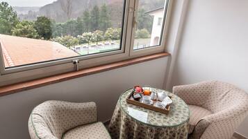 Una stanza piccola con due poltrone decorate, un tavolino in vetro con un vassoio di cibi per la colazione e una finestra che si affaccia su un giardino e degli edifici.