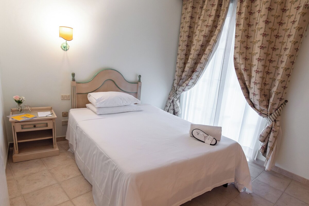A neatly made bed with white linens, a bedside table with a lamp, and a window with floral curtains.