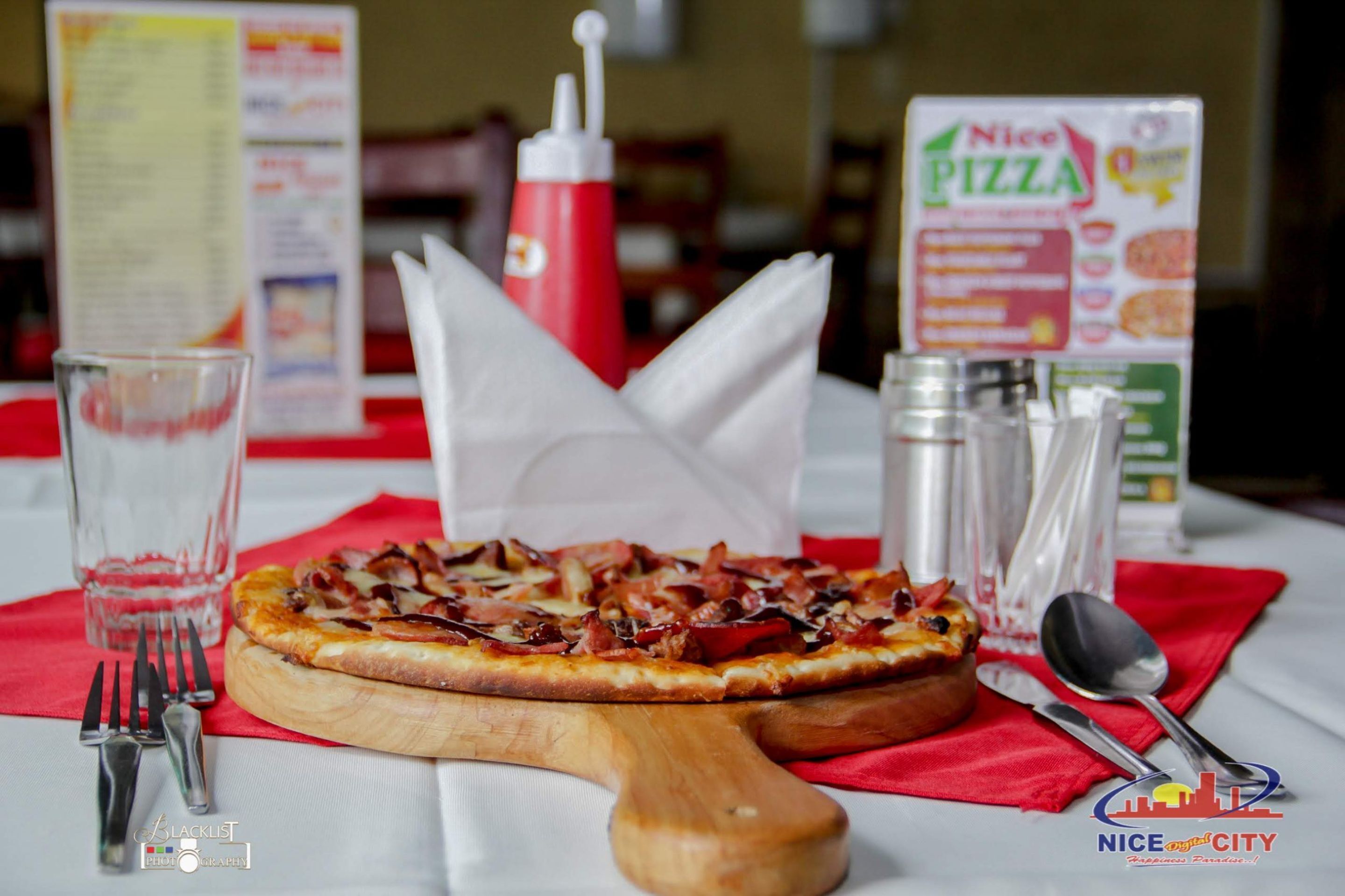 Pizza auf einem Holzbrett mit einer roten Serviette, Besteck und einer Speisekarte im Hintergrund.