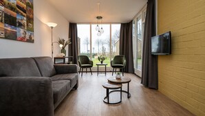 Un salon moderne comprenant un canapé, deux fauteuils, une petite table, un téléviseur fixé au mur et de grandes fenêtres avec des rideaux.