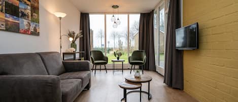 A modern living room with a sofa, two chairs, a small table, a TV mounted on the wall, and large windows with curtains.