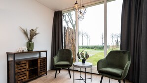Une pièce comprenant deux fauteuils verts, une petite table ronde, une console en bois ornée d’un vase et d’objets décoratifs, et une grande fenêtre donnant sur un jardin.
