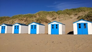 Une rangée de cabanes de plage numérotées de 72 à 76, avec des portes bleues et des murs blancs, située sur une plage de sable avec des dunes en arrière-plan.