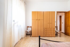 A bedroom with a bed, a wooden wardrobe, a chair, and a window with sheer curtains.