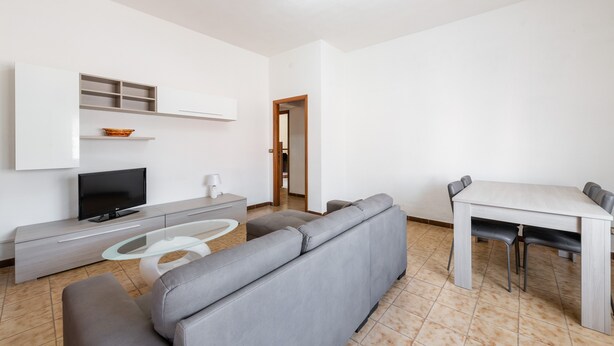 Ein modernes Wohnzimmer mit grauem Sofa, einem Glastisch, einem Flachbildfernseher, einer weißen Kommode und einem Esstisch mit Stühlen.