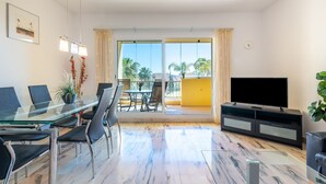 A modern living room with a glass table, black chairs, a flat-screen TV, and a balcony with a view of palm trees. - Modern Apartment 'In Jardin Botanico' with Sea View, Pool, Wi-Fi, Garden & Terrace (La Cala de Mijas)
