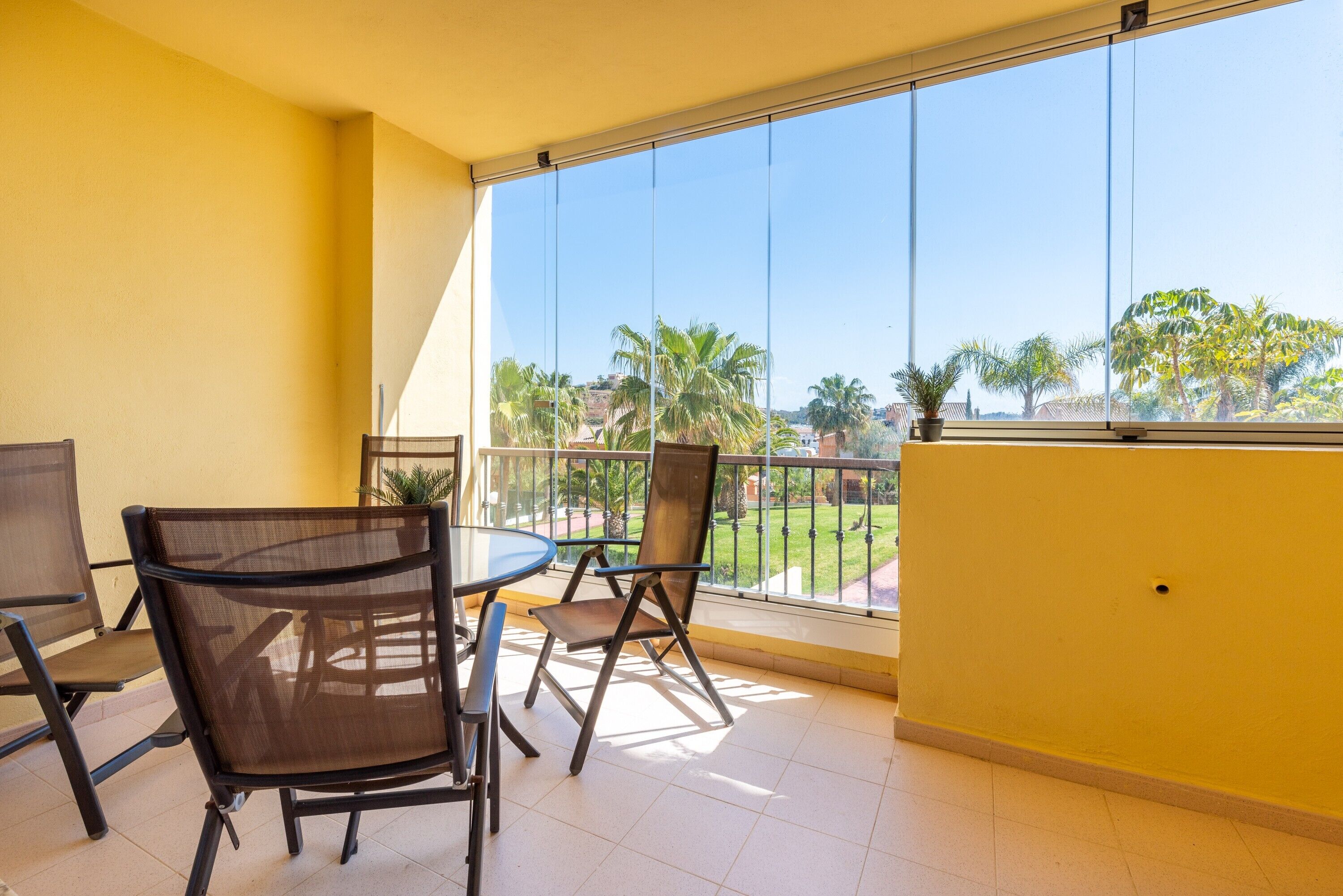 A balcony with a glass railing, a table with chairs, and a view of palm trees and buildings.