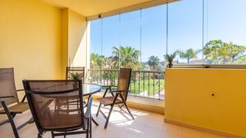A balcony with a glass railing, a table with chairs, and a view of palm trees and buildings.