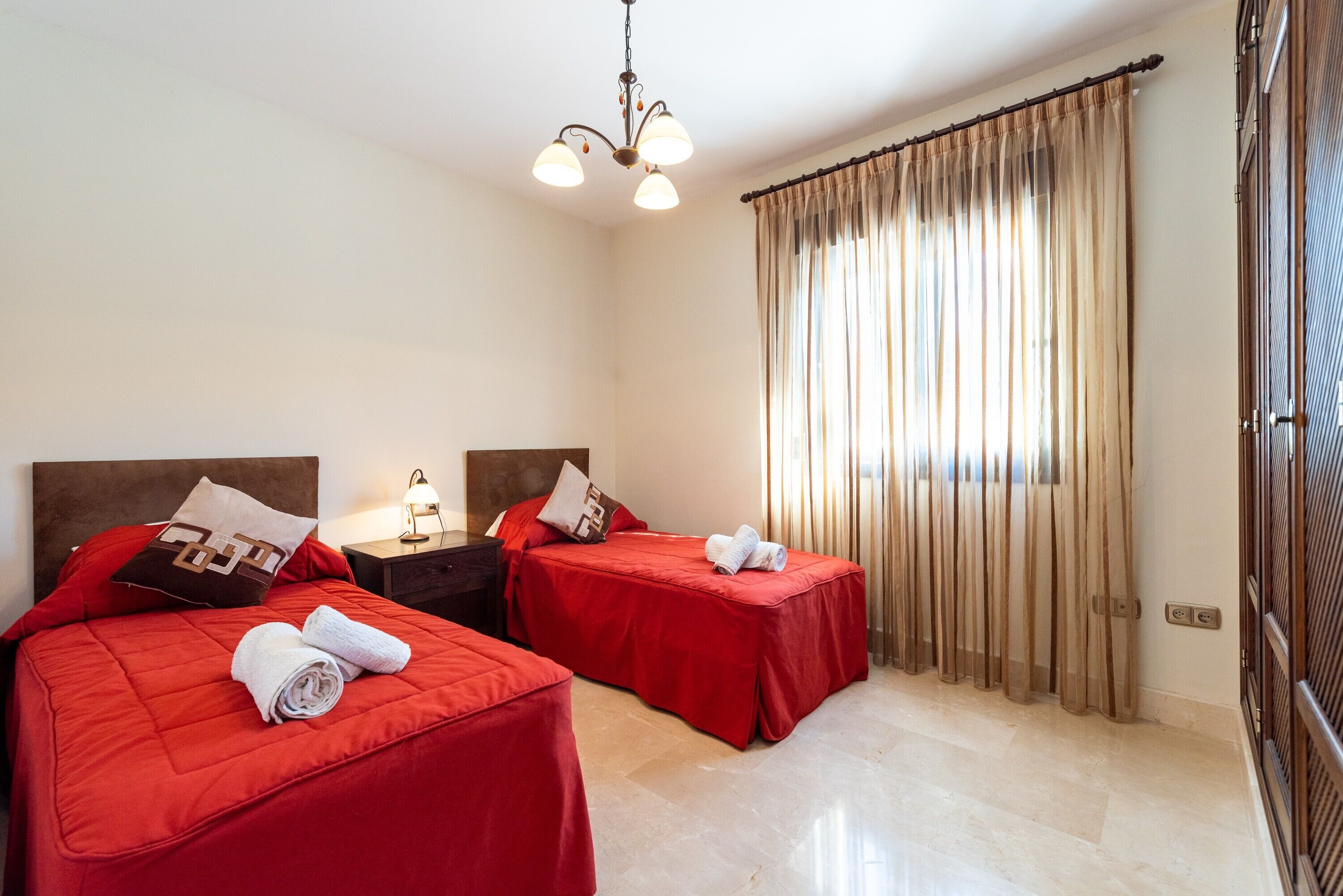 A hotel room with two beds, a chandelier, a nightstand, and a window with curtains.