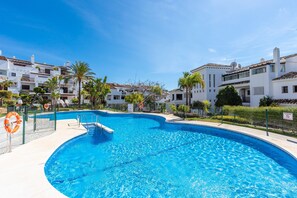 A swimming pool surrounded by white buildings with balconies, palm trees, and a clear blue sky.
