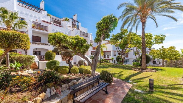 A courtyard with a bench, palm trees, and a white building with balconies. - Apartment "Finca San Antonio Holiday Rental" with Pool, Terrace & Wi-Fi (Mijas Pueblo)