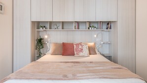 Un dormitorio con una cama, una mesita de noche, una estantería con libros y plantas, y una lámpara de pared.