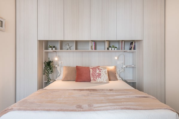 Une chambre à coucher comprenant un lit, une table de chevet, une étagère avec des livres et des plantes, et un luminaire fixé au mur.