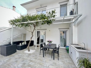 Une terrasse avec une table et des chaises, un petit arbre et un bâtiment blanc doté d’un balcon.