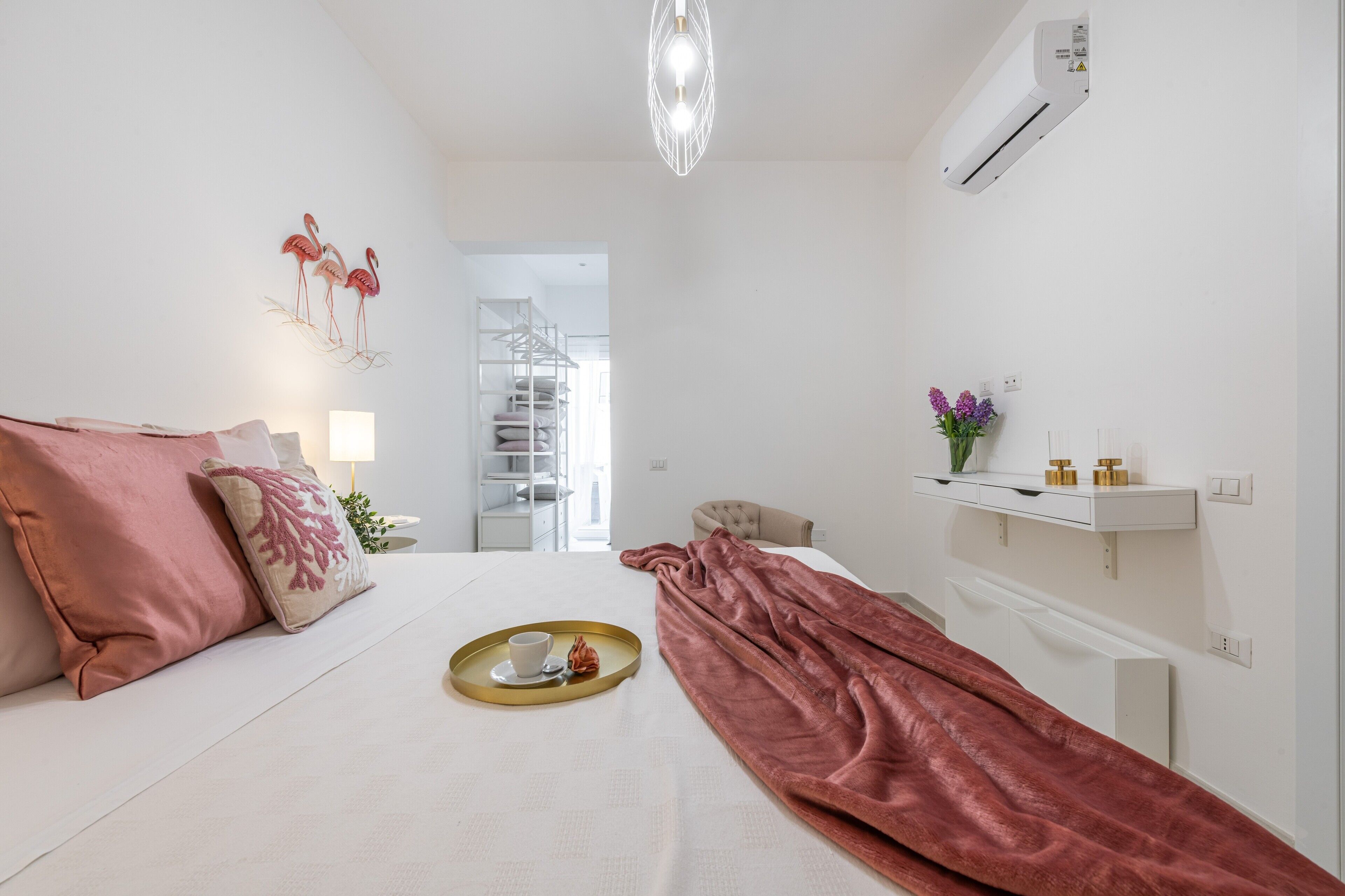 Una camera d'albergo con un letto, un comodino con vassoio, una lampada, uno scaffale con articoli da toilette e una scala sullo sfondo.