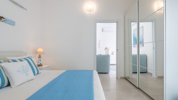 A bedroom with a bed, bedside table, lamp, and a view into another room through a door. - Pet-Friendly Apartment 'La Casa dei Naviganti' with Courtyard and Wi-Fi (Villasimius)