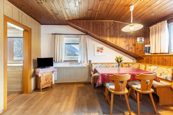 A cozy cabin interior with a wooden ceiling, a dining table with chairs, a sofa, a radiator, and a television on a wooden cabinet.