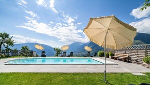 Une piscine entourée de chaises longues et de parasols, délimitée par une clôture en bois, avec des montagnes en toile de fond.
