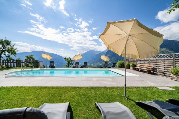 A swimming pool with lounge chairs and umbrellas, surrounded by a wooden fence and mountains in the background.