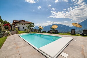 A rectangular swimming pool with a white edge, surrounded by a stone patio. There are four lounge chairs and two umbrellas. A white house with a brown roof is visible in the background.
