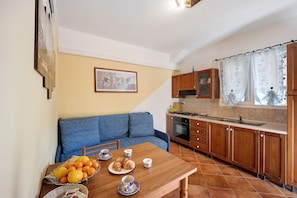A compact kitchen with wooden cabinets, a dining table with fruits and pastries, and a blue sofa.