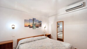 Een slaapkamer met een bed, een wandairconditioner, een nachtkastje, een spiegel en een stadsgezicht schilderij.