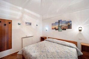 A bedroom with a bed, a wooden wardrobe, a radiator, a nightstand, a wall-mounted light, and a cityscape painting.
