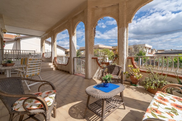 Une terrasse couverte, aménagée avec des meubles en osier, des plantes en pot et offrant une vue sur le quartier.